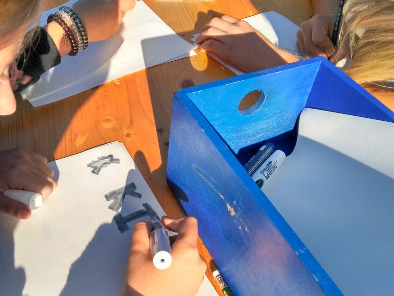 KI generiert: Das Bild zeigt eine Gruppe von Personen, die an einem Holztisch sitzen und auf Papier zeichnen oder schreiben. Der Hauptinhalt des Bildes ist eine kreative oder künstlerische Aktivität, bei der verschiedene Werkzeuge wie Stifte, Marker und ein blauer Behälter mit Materialien verwendet werden.