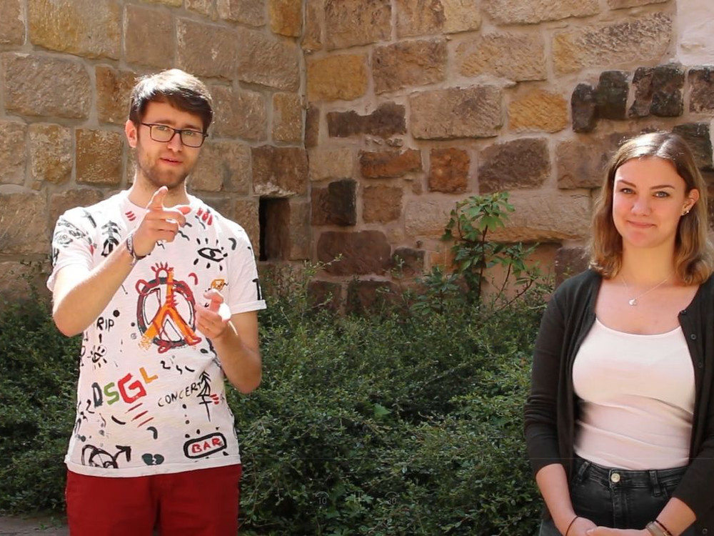 KI generiert: Das Bild zeigt zwei Personen, einen Mann und eine Frau, die vor einer alten Steinmauer und einem Busch stehen. Der Mann trägt ein bunt bedrucktes T-Shirt und hält einen Gegenstand in der Hand, während die Frau ein weißes Oberteil und eine dunkle Jacke trägt.