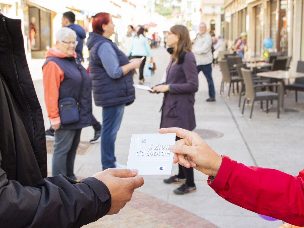 KI generiert: Das Bild zeigt eine Person in einem roten Mantel, die einer anderen Person ein Stück Papier mit der Aufschrift "#ZIVILCOURAGE" überreicht. Im Hintergrund sind mehrere Menschen auf einer belebten Straße zu sehen, einige von ihnen scheinen sich zu unterhalten.