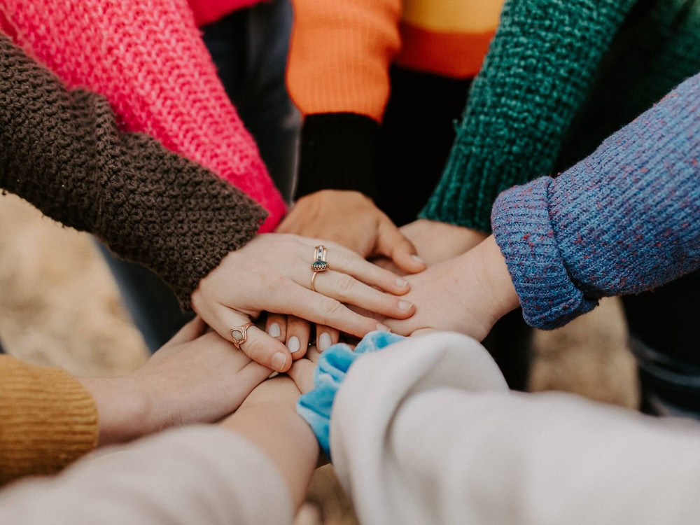 KI generiert: Das Bild zeigt mehrere Hände, die sich in der Mitte übereinanderlegen, was Symbol für Zusammenhalt und Teamgeist steht. Die Personen tragen verschiedene Kleidungsstücke, darunter ein blauer Pullover und ein brauner Mantel.