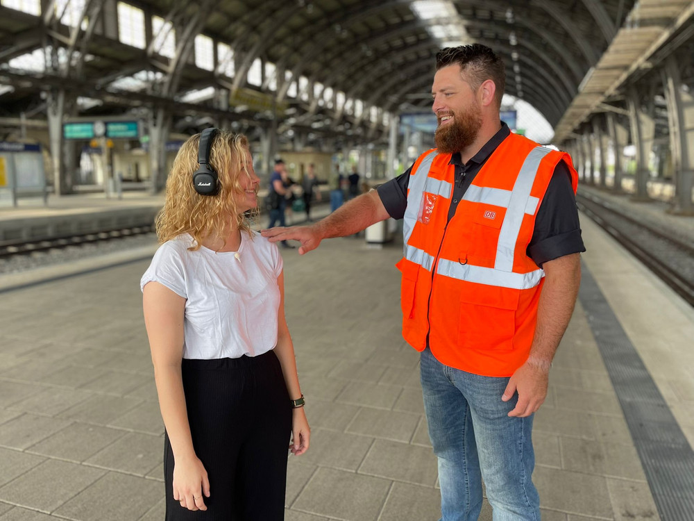 KI generiert: Das Bild zeigt eine Frau mit Kopfhörern und einen Mann in einer orangefarbenen Warnweste, die sich auf einem Bahnsteig unterhalten. Im Hintergrund sind weitere Personen und die Architektur eines Bahnhofs zu erkennen.