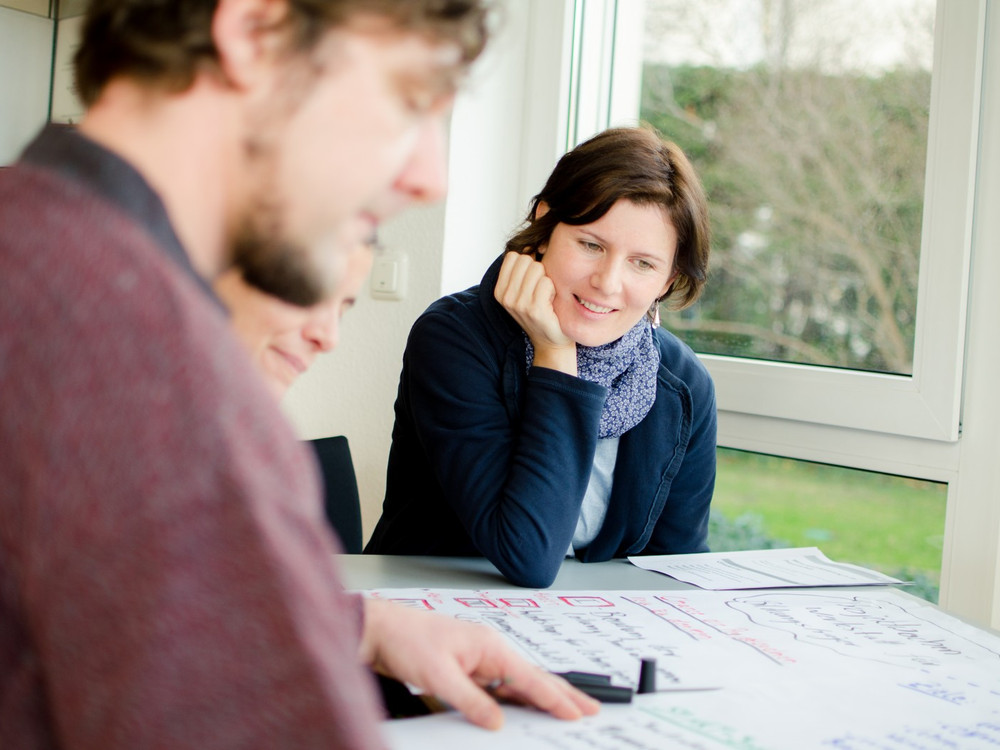 KI generiert: Das Bild zeigt zwei Personen, die an einem Tisch sitzen und sich auf ein Blatt Papier konzentrieren, das mit Notizen und Zeichnungen bedeckt ist. Der Hauptinhalt des Bildes ist eine Arbeits- oder Brainstorming-Sitzung.