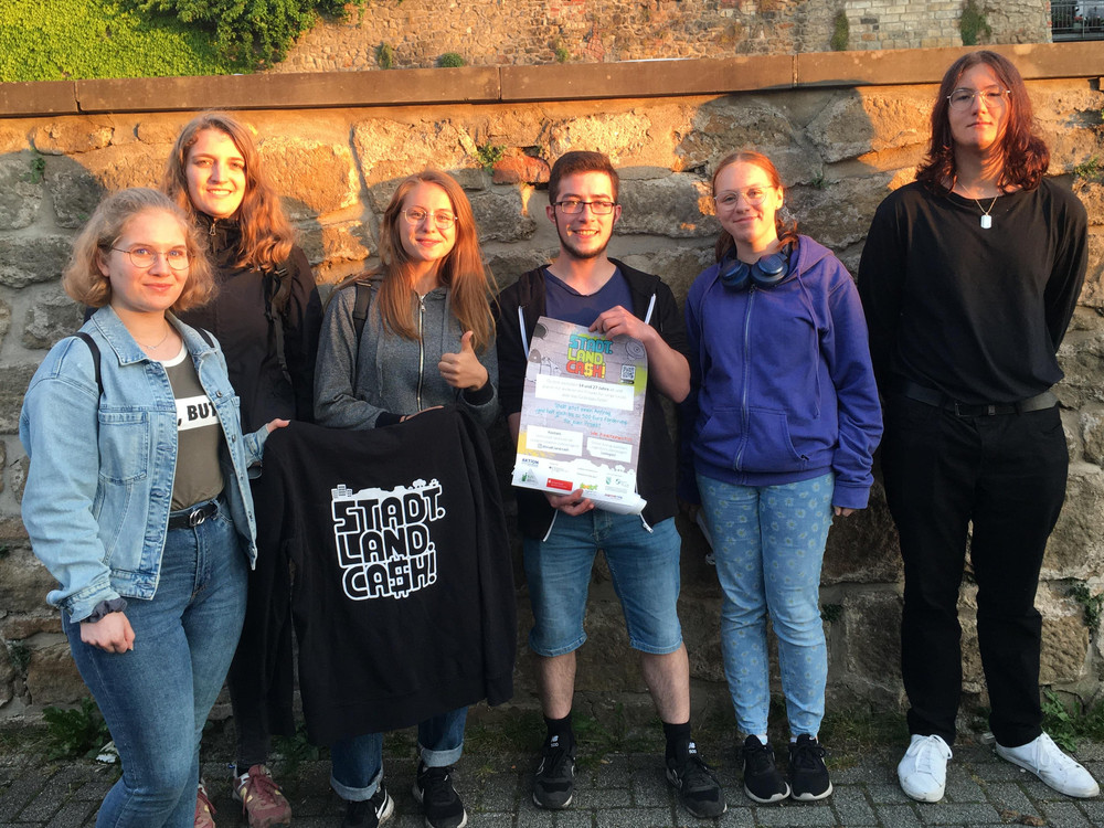 KI generiert: Das Bild zeigt eine Gruppe von sechs Personen, die vor einer alten Steinmauer stehen und in die Kamera lächeln. Eine Person in der Mitte hält ein Zertifikat oder eine Urkunde in den Händen.