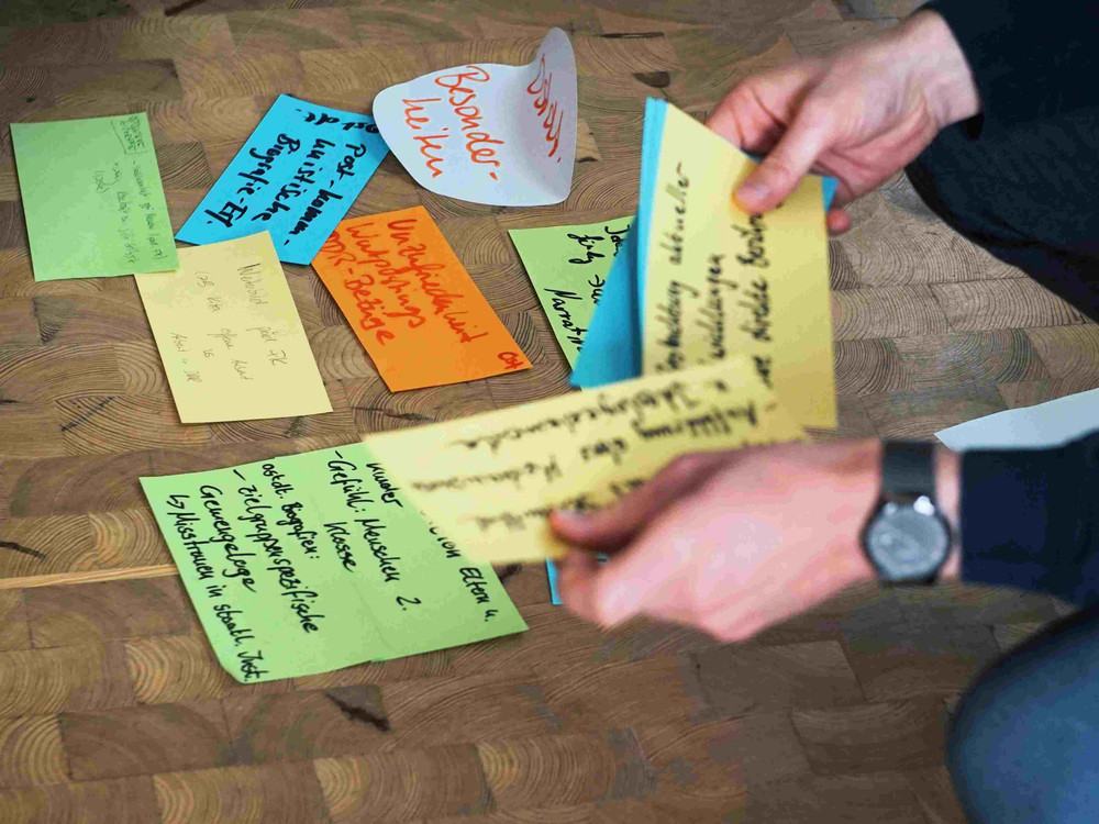 KI generiert: Das Bild zeigt eine Gruppe von Personen, die in einem hellen Raum mit großen Fenstern an einer Besprechung oder einem Workshop teilnehmen. Ein Mann steht in der Mitte und präsentiert etwas, während die anderen auf Stühlen sitzen und zuhören, wobei an den Wänden verschiedene Notizen und Diagramme an Whiteboards hängen.