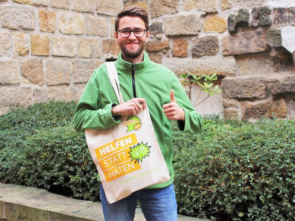 KI generiert: Das Bild zeigt einen lächelnden Mann mit Brille, der einen grünen Pullover trägt und einen Beutel mit der Aufschrift "HELFEN STATT HATEN" in der Hand hält, während er ein Daumen-hoch-Zeichen gibt. Der Hintergrund besteht aus einer Steinmauer und grünen Büschen.