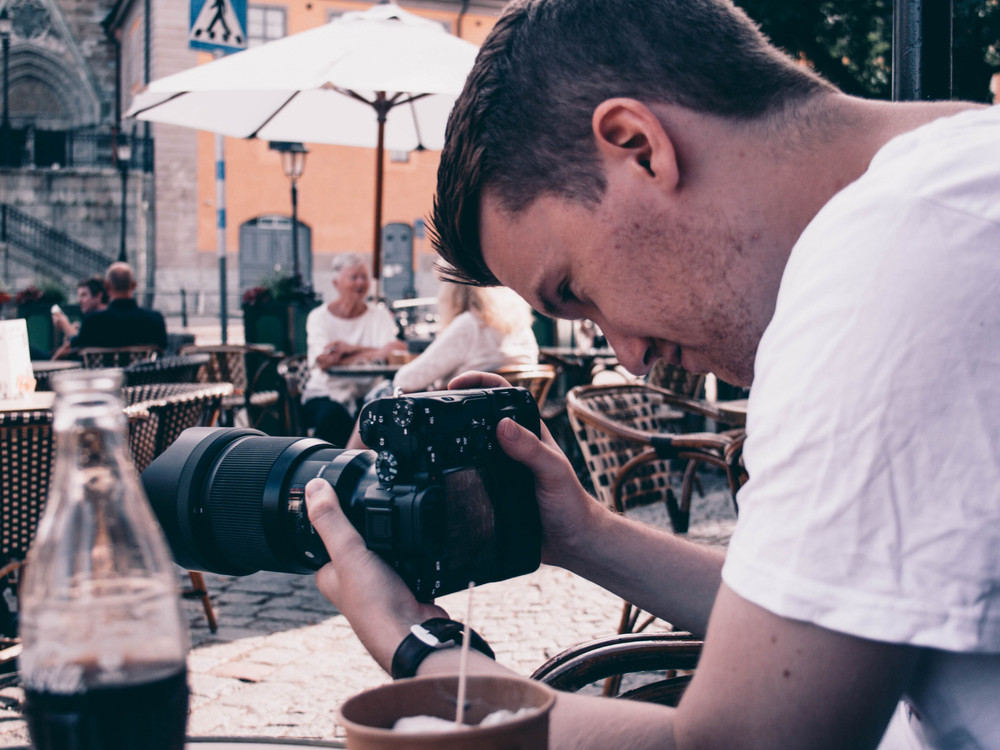 KI generiert: Das Bild zeigt einen Mann, der sich über einen Tisch beugt und eine Kamera mit einem großen Teleobjektiv bedient. Im Vordergrund steht ein Glas mit einem dunklen Getränk, möglicherweise Cola, und im Hintergrund sind Menschen an Tischen in einem Café oder einer öffentlichen Platzsituation zu sehen. Der Hauptinhalt des Bildes ist die Fotografie und die entspannte Atmosphäre eines öffentlichen Ortes.