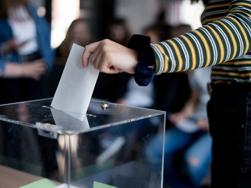 KI generiert: Das Bild zeigt eine Person, die einen Wahlzettel in eine Wahlurne wirft. Der Hauptinhalt des Bildes ist die Darstellung des Wahlvorgangs.