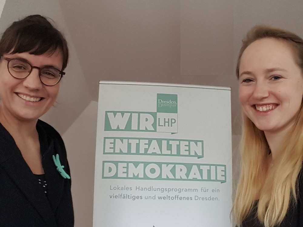 KI generiert: Das Bild zeigt zwei lächelnde Frauen, die neben einem Plakat mit der Aufschrift "Wir entfalten Demokratie" stehen. Das Plakat wirbt für ein lokales Handlungsprogramm für ein vielfältiges und weltoffenes Dresden.