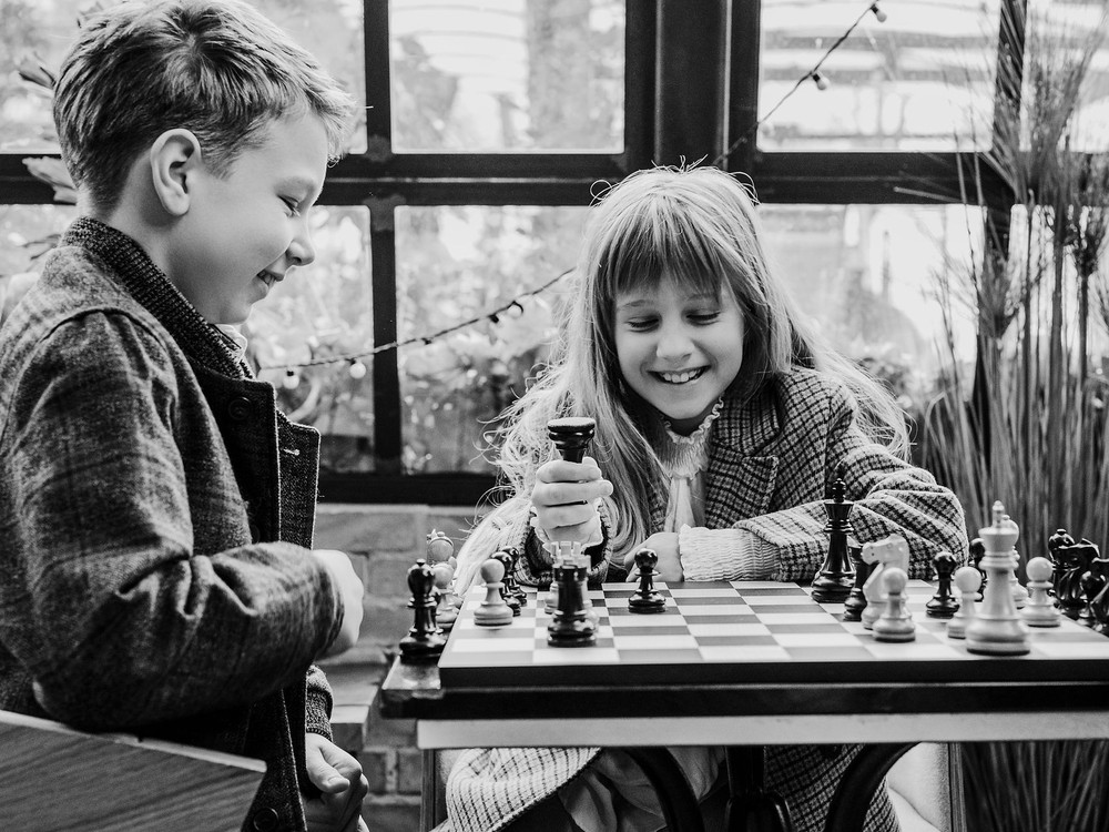 KI generiert: Das Bild zeigt zwei Personen, die an einem Tisch sitzen und Schach spielen, wobei eine junge Person im Vordergrund einen Zug macht. Der Hauptinhalt des Bildes ist das Schachspiel zwischen den beiden Personen.