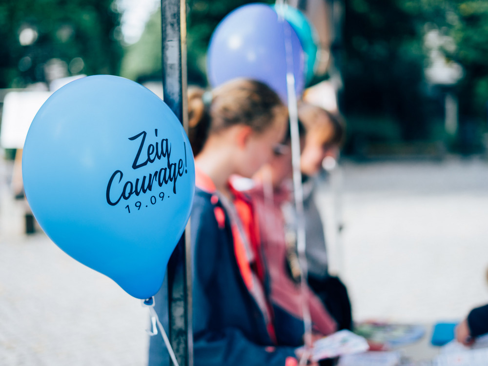 KI generiert: Das Bild zeigt einen blauen Luftballon mit der Aufschrift "Zeig Courage! 19.09." im Vordergrund, während im Hintergrund unscharf einige Personen zu sehen sind, die möglicherweise an einer Veranstaltung teilnehmen. Der Hauptinhalt des Bildes ist der Luftballon mit der Botschaft, die auf eine mutige Handlung oder ein Event hinweist.