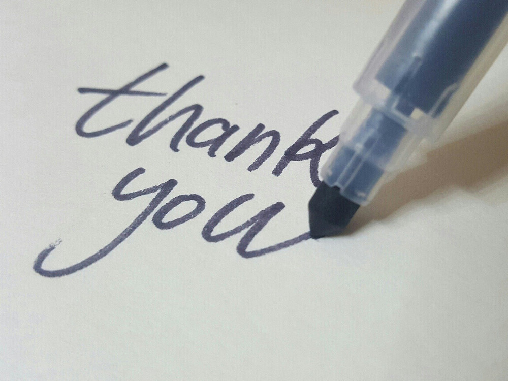 KI generiert: Das Bild zeigt einen Ausschnitt eines handgeschriebenen "thank you" auf Papier mit einem blauen Stift, der gerade den Text schreibt. Der Hauptinhalt des Bildes ist der Ausdruck von Dankbarkeit.