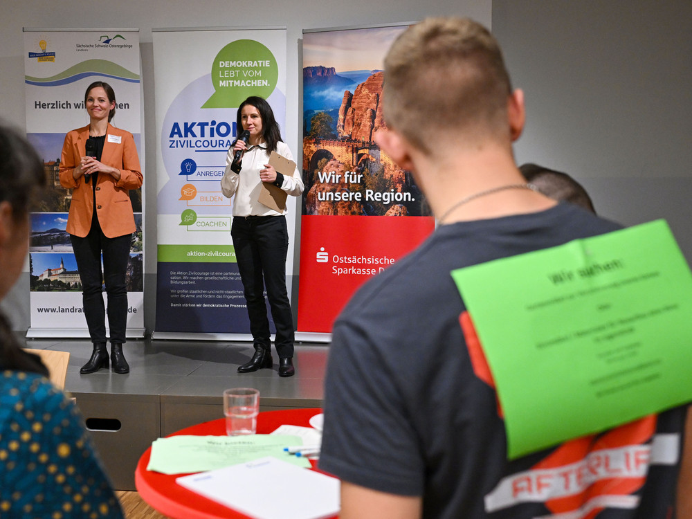 KI generiert: Das Bild zeigt eine belebte Indoor-Veranstaltung mit vielen Menschen, die sich an verschiedenen Ständen und Tischen unterhalten. Der Hauptinhalt des Bildes ist eine Netzwerk- oder Informationsveranstaltung, bei der die Teilnehmer in einer entspannten Atmosphäre interagieren.