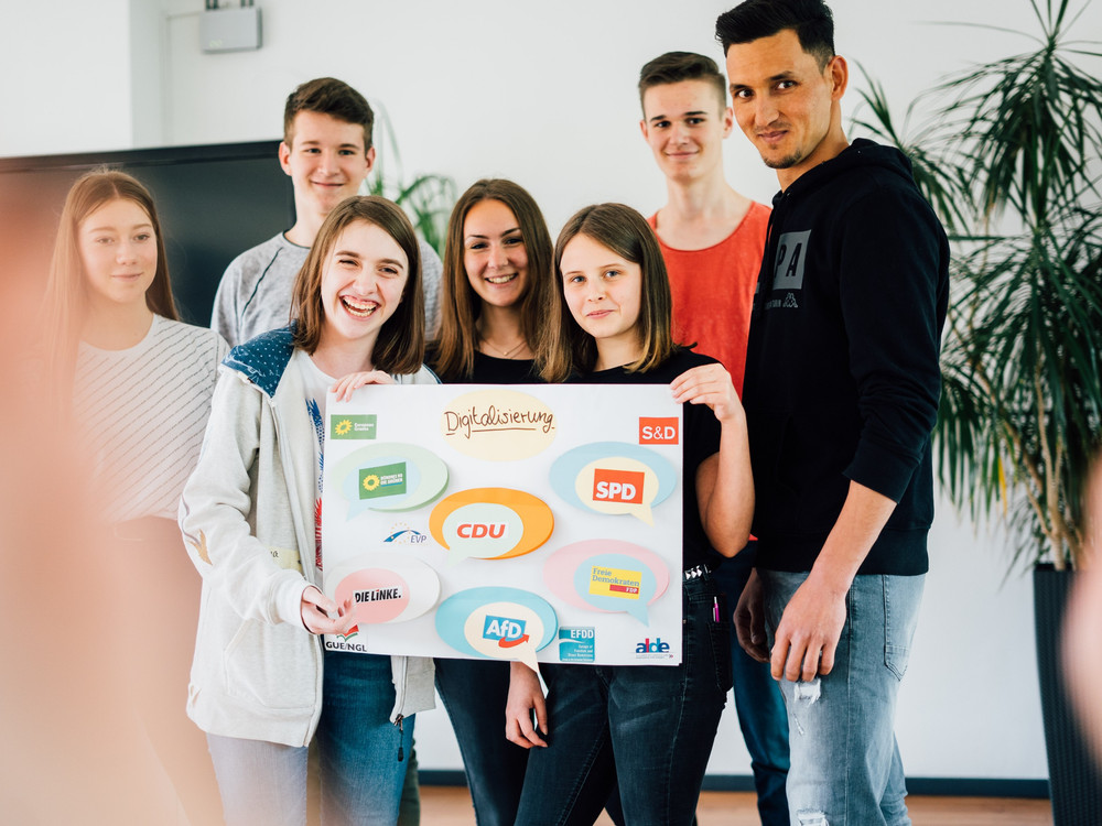 KI generiert: Das Bild zeigt eine Gruppe von sieben Personen, die gemeinsam ein Plakat mit dem Titel "Digitalisierung" und verschiedenen Logos halten. Der Hauptinhalt des Bildes ist die Darstellung einer Teamarbeit oder eines Projekts im Bereich Digitalisierung.