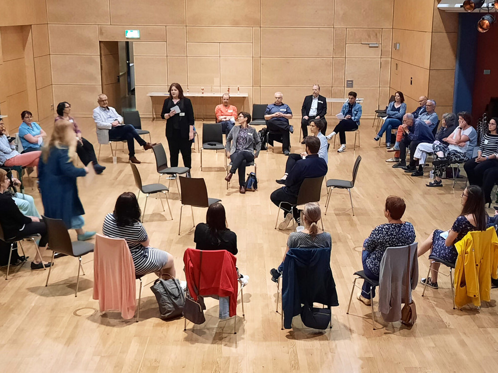 KI generiert: Das Bild zeigt eine Gruppe von Menschen, die in einem Kreis in einem Raum mit Holzvertäfelung sitzen und sich anscheinend in einer Diskussion oder einem Workshop befinden. Der Hauptinhalt des Bildes ist eine interaktive Gruppenaktivität oder ein Meeting.