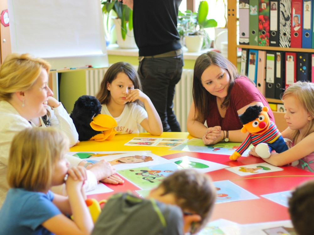 KI generiert: Das Bild zeigt eine Gruppe von Kindern und Erwachsenen, die um einen bunten Tisch sitzen und scheinbar an einer Aktivität oder einem Spiel teilnehmen. Der Hauptinhalt des Bildes ist eine pädagogische oder spielerische Interaktion in einer Kindertagesstätte oder einem Klassenzimmer.