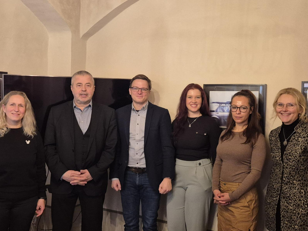 KI generiert: Das Bild zeigt eine Gruppe von sechs Personen, die nebeneinander in einem Innenraum stehen und in die Kamera lächeln. Im Hintergrund sind zwei Bildschirme oder gerahmte Bilder an der Wand zu sehen.