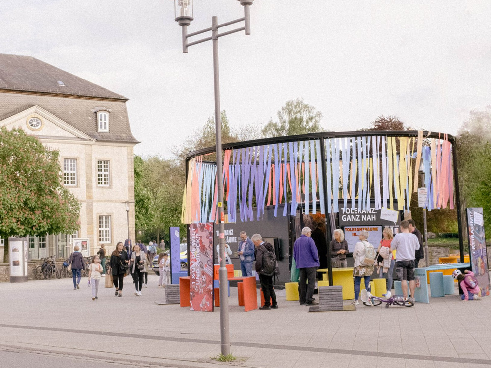 KI generiert: Das Bild zeigt eine nächtliche Szene mit einem beleuchteten Container, der als Ausstellungsraum genutzt wird und auf dem "WE'RE OPEN!" steht. Im Hintergrund sind bunte Bänder und weitere Ausstellungsstände zu sehen, die auf eine kulturelle oder künstlerische Veranstaltung hinweisen.