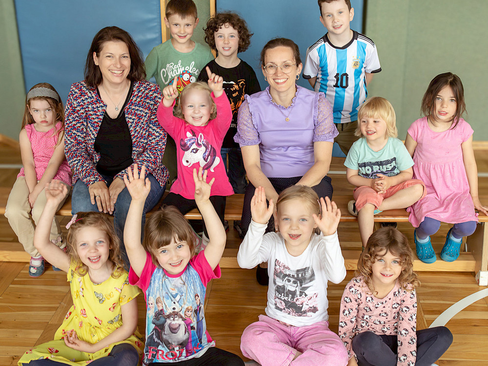 KI generiert: Das Bild zeigt eine Gruppe fröhlicher Kinder, die auf einer Holzbank sitzen und ihre Hände in die Luft strecken. Der Hauptinhalt des Bildes ist die Darstellung von mutigen Kindern, die Unterstützung benötigen, wie der Text "Couragierte Kinder - Wir brauchen eure Unterstützung" andeutet.