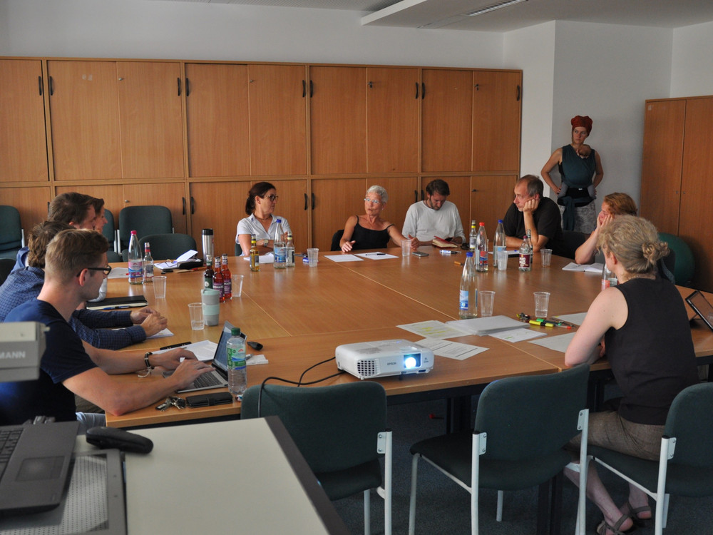KI generiert: Das Bild zeigt eine Gruppe von Personen, die in einem Besprechungsraum an einem großen ovalen Tisch sitzen und sich in einer Besprechung oder einem Meeting befinden. Der Hauptinhalt des Bildes ist eine formelle Zusammenkunft oder Diskussion in einem Büroumfeld.