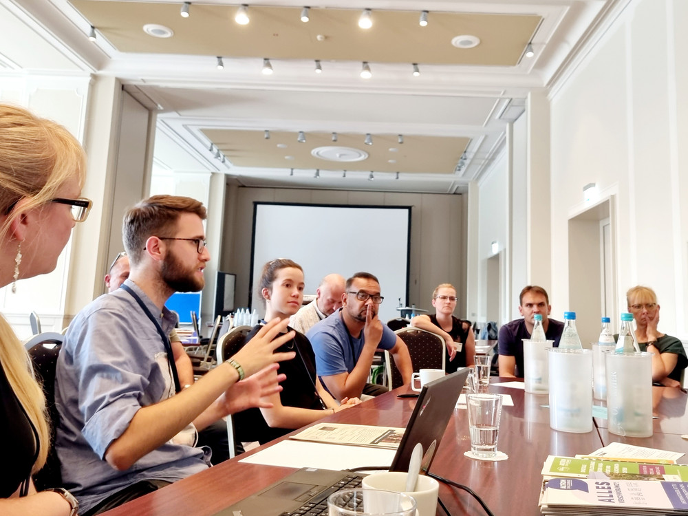 KI generiert: Das Bild zeigt eine Gruppe von Personen, die an einem Tisch sitzen und auf Tablets oder Laptops arbeiten. Der Hauptinhalt des Bildes ist eine Lernsituation oder ein Workshop, bei dem digitale Geräte genutzt werden.