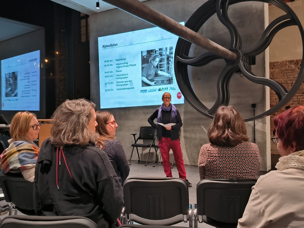 KI generiert: Das Bild zeigt eine Person, die vor einer Gruppe von Zuhörern spricht, während eine Präsentation auf einem Bildschirm im Hintergrund läuft. Der Hauptinhalt des Bildes ist eine Vortrags- oder Diskussionsveranstaltung in einem industriell gestalteten Raum.