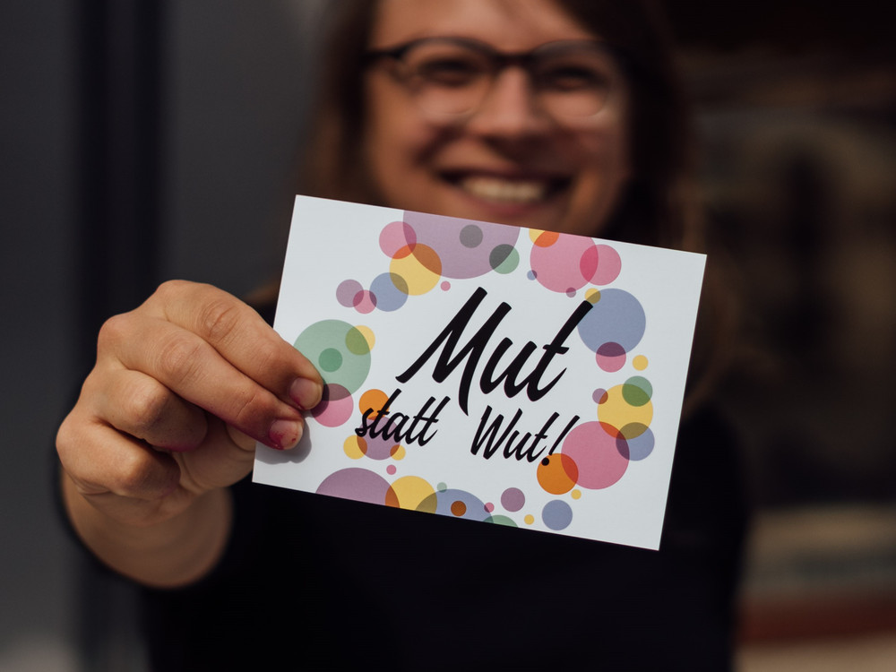KI generiert: Das Bild zeigt eine Person, die ein weißes Kartenblatt mit bunten Kreisen und der Aufschrift "Mut statt Wut!" in der Hand hält. Der Hauptinhalt des Bildes ist die Botschaft auf der Karte, die Mut und Positivität fördert.