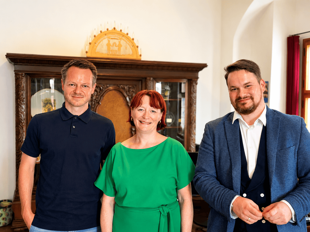 KI generiert: Das Bild zeigt drei Personen, die in einem Raum mit antiker Einrichtung stehen und in die Kamera lächeln. Der Hauptinhalt des Bildes ist die Darstellung der drei Personen vor einem alten Schrank mit einer goldenen Krone darauf.