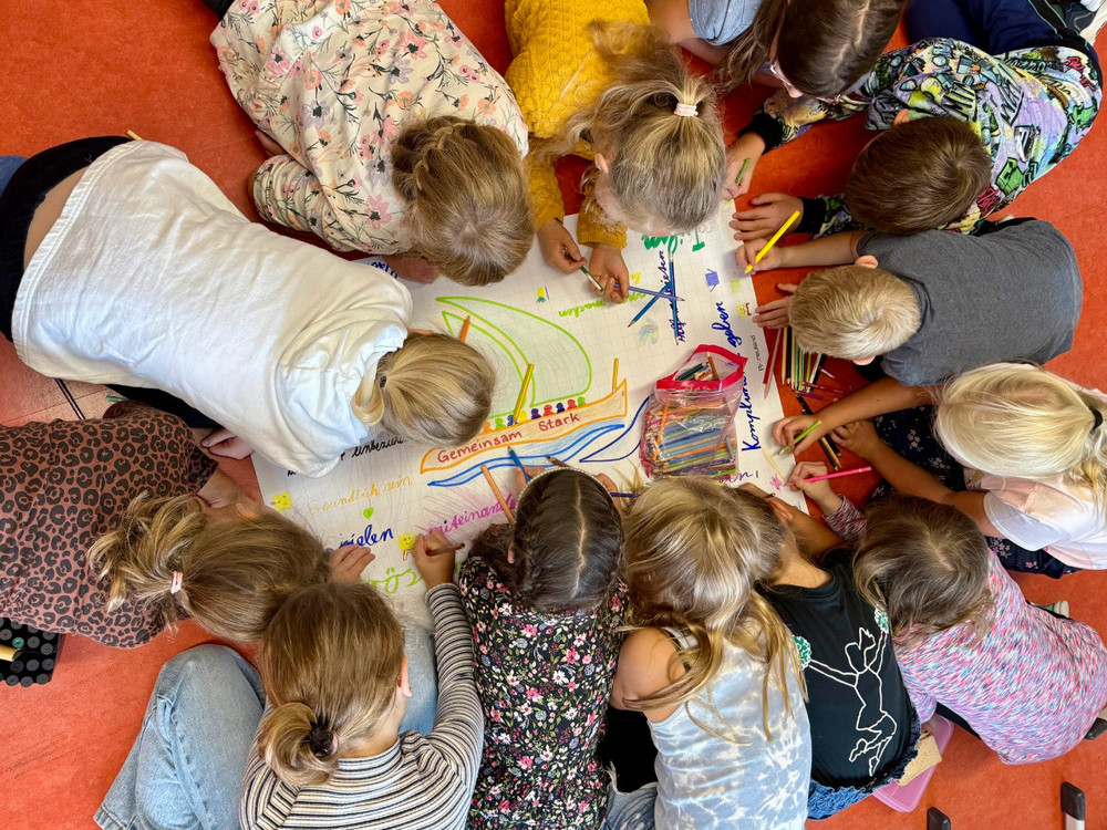 KI generiert: Das Bild zeigt eine Gruppe von Kindern, die gemeinsam auf einem großen Blatt Papier malen und zeichnen. Der Hauptinhalt des Bildes ist die kreative Zusammenarbeit und das gemeinsame Gestalten der Kinder.