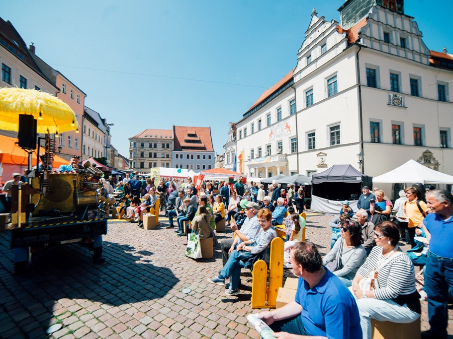 KI generiert: Das Bild zeigt eine belebte Straßenveranstaltung mit vielen Menschen, die auf Bänken sitzen und sich unterhalten, während im Hintergrund historische Gebäude und Marktstände zu sehen sind. Im Vordergrund steht ein altes, gelbes Fahrzeug mit einem großen gelben Schirm.