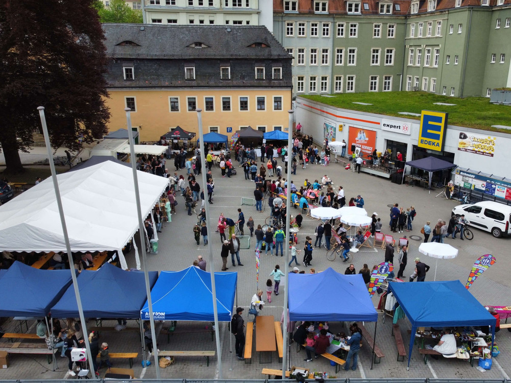 KI generiert: Das Bild zeigt eine belebte Outdoor-Veranstaltung mit vielen Menschen, die zwischen Ständen, Zelten und einem Gebäude mit Werbebannern umherlaufen. Im Vordergrund sind weiße Sonnenschirme und ein Fahrrad zu sehen, während im Hintergrund weitere Besucher und Aussteller zu erkennen sind.