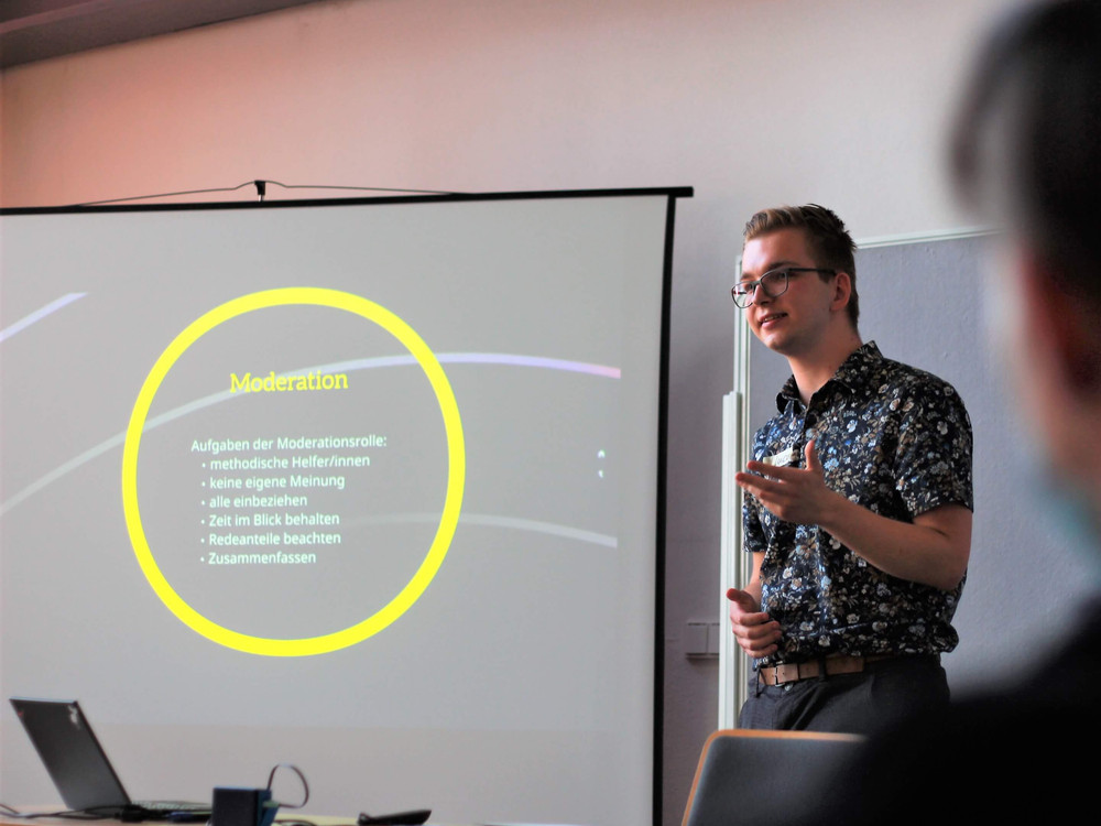 KI generiert: Das Bild zeigt einen Mann, der vor einem Projektor steht und eine Präsentation hält. Der Hauptinhalt der Präsentation scheint das Thema "Moderation" zu sein, wobei spezifische Punkte wie die Aufgaben der Moderationsrolle und methodische Hilfsmittel erwähnt werden.