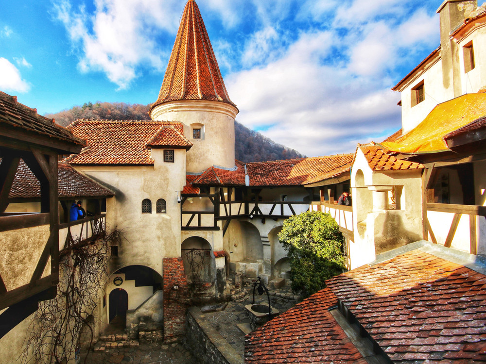 KI generiert: Das Bild zeigt eine malerische Burg mit roten Ziegeldächern und einem runden Turm, umgeben von einer bergigen Landschaft. Im Vordergrund sind Holzkonstruktionen und ein Innenhof zu sehen, in dem sich einige Personen aufhalten.