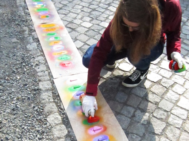 KI generiert: Das Bild zeigt eine Person, die auf einem Gehweg mit Sprühdosen bunte Graffiti auf eine lange Papierbahn sprüht. Der Hauptinhalt des Bildes ist das Graffiti-Sprühen als künstlerische Aktivität.