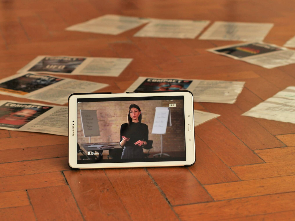 KI generiert: Das Bild zeigt drei Personen, die in einem Raum stehen und jeweils ein Blatt Papier in den Händen halten. Es scheint sich um eine Besprechung oder Präsentation zu handeln, da im Hintergrund eine Projektionsleinwand mit Text zu sehen ist.