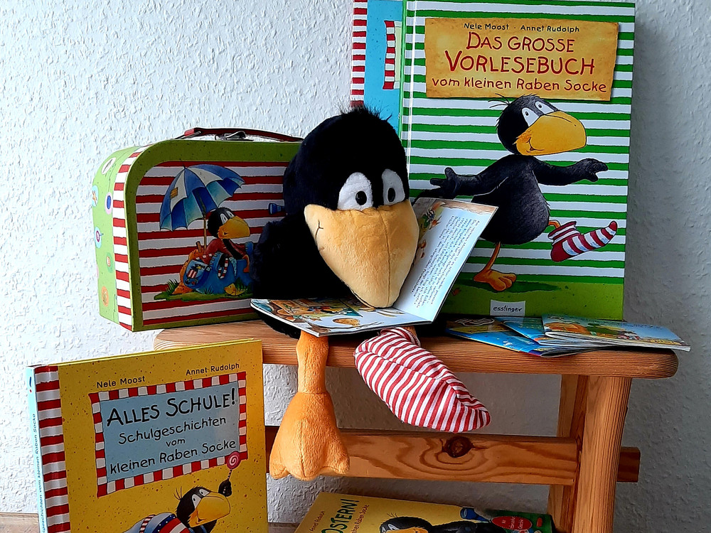 KI generiert: Das Bild zeigt ein Stofftier in Form eines Raben, das ein Buch in den Flügeln hält. Im Hintergrund ist eine Kindertragtasche mit bunten Motiven zu sehen.