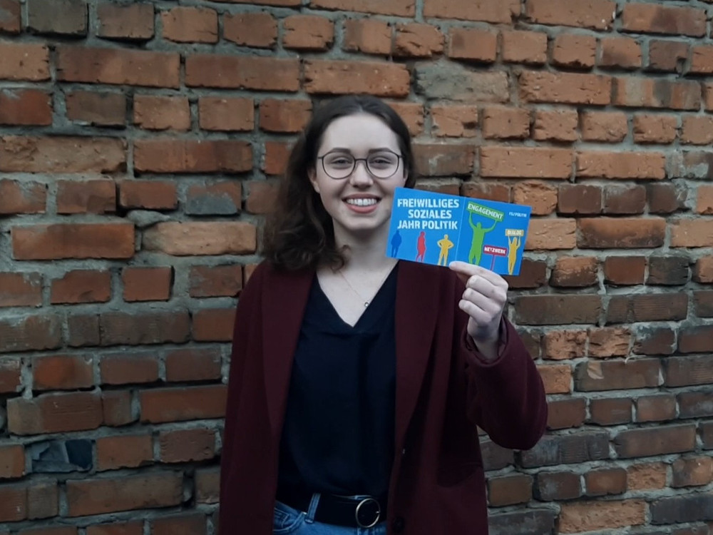 KI generiert: Das Bild zeigt eine lächelnde Person mit Brille, die vor einer Ziegelsteinmauer steht und ein Buch mit dem Titel "Freiwilliges Soziales Jahr Politik" in der Hand hält. Der Hauptinhalt des Bildes ist die Präsentation des Buches.