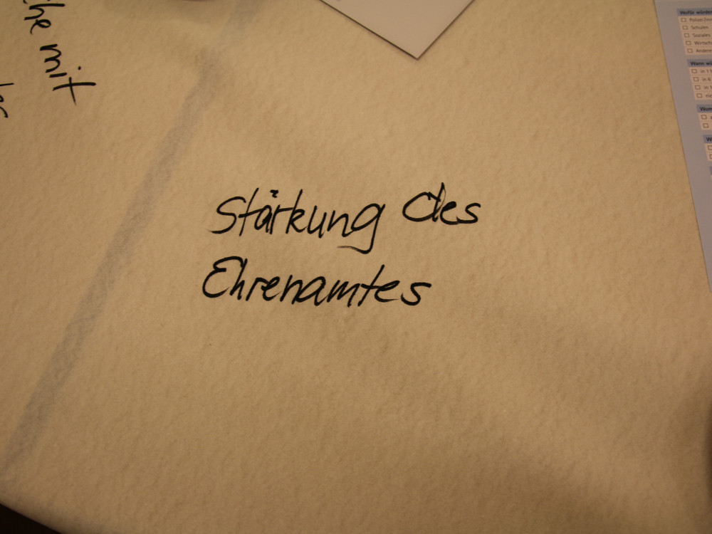 KI generiert: Das Bild zeigt eine Tischdecke mit der handschriftlichen Aufschrift "Stärkung des Ehrenamtes". Im Hintergrund sind weitere handschriftliche Notizen und ein Blatt Papier zu erkennen.