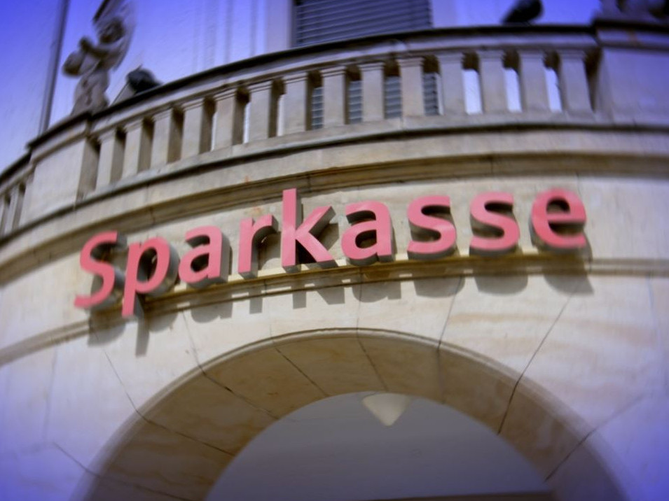 KI generiert: Das Bild zeigt die Fassade eines Gebäudes mit dem leuchtend roten Schriftzug "Sparkasse" darüber. Der Hauptinhalt des Bildes ist das Logo der Sparkasse, einer bekannten Bank in Deutschland.