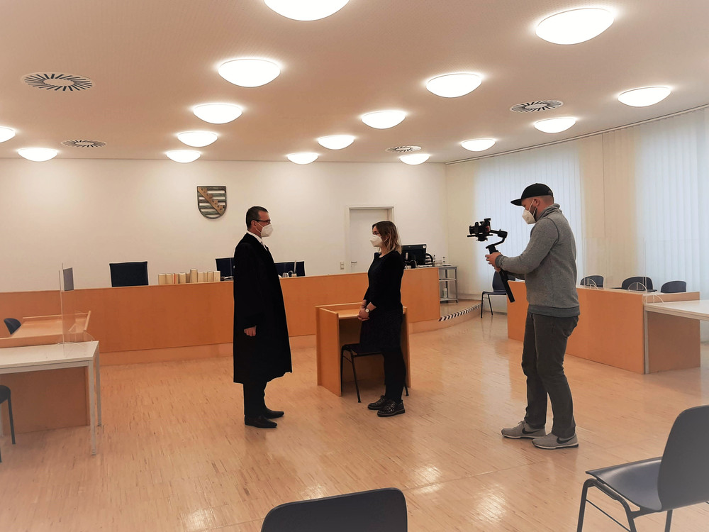 KI generiert: Das Bild zeigt einen Gerichtssaal, in dem eine Person in Robe und eine weitere Person vor einem Richtertisch stehen, während eine dritte Person mit einer Kamera die Szene filmt. Der Hauptinhalt des Bildes ist eine gerichtliche Verhandlung oder ein gerichtliches Verfahren, das aufgezeichnet wird.