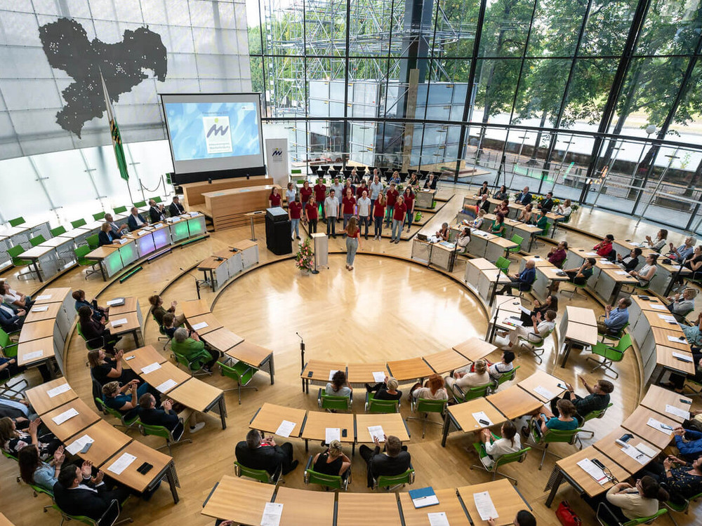 KI generiert: Das Bild zeigt eine Gruppe von Menschen, die in einem hellen Raum stehen und Blumensträuße sowie Urkunden oder Preise in den Händen halten. Es scheint sich um eine feierliche Veranstaltung oder eine Preisverleihung zu handeln.
