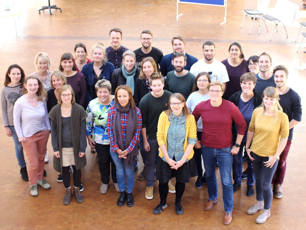 KI generiert: Das Bild zeigt eine Gruppe von etwa 25 Personen, die in einem hellen Innenraum mit Holzfußboden und einigen Stühlen im Hintergrund posieren. Der Hauptinhalt des Bildes ist die Gruppenfotografie, die eine vielfältige Gruppe von Menschen in lockerer, freundlicher Atmosphäre zeigt.