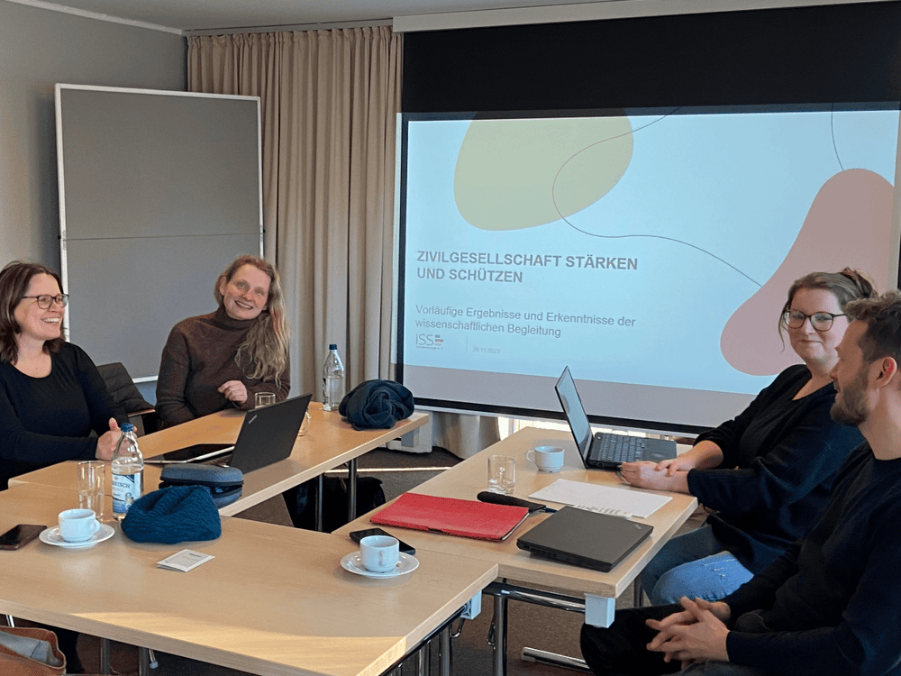 KI generiert: Das Bild zeigt eine Gruppe von vier Personen, die an einem Tisch sitzen und an Laptops arbeiten, während eine Präsentation im Hintergrund auf einem Bildschirm läuft. Der Hauptinhalt des Bildes scheint eine Arbeitsbesprechung oder ein Workshop zu sein, bei dem die Teilnehmer gemeinsam an einem Projekt arbeiten.