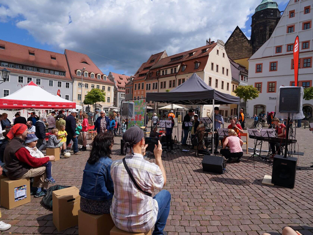 KI generiert: Das Bild zeigt eine belebte Straßenveranstaltung mit Menschen, die sich um Stände und eine kleine Bühne mit Musikern versammeln. Der Hauptinhalt ist ein öffentliches Fest oder Markt in einer historischen Stadtkulisse.