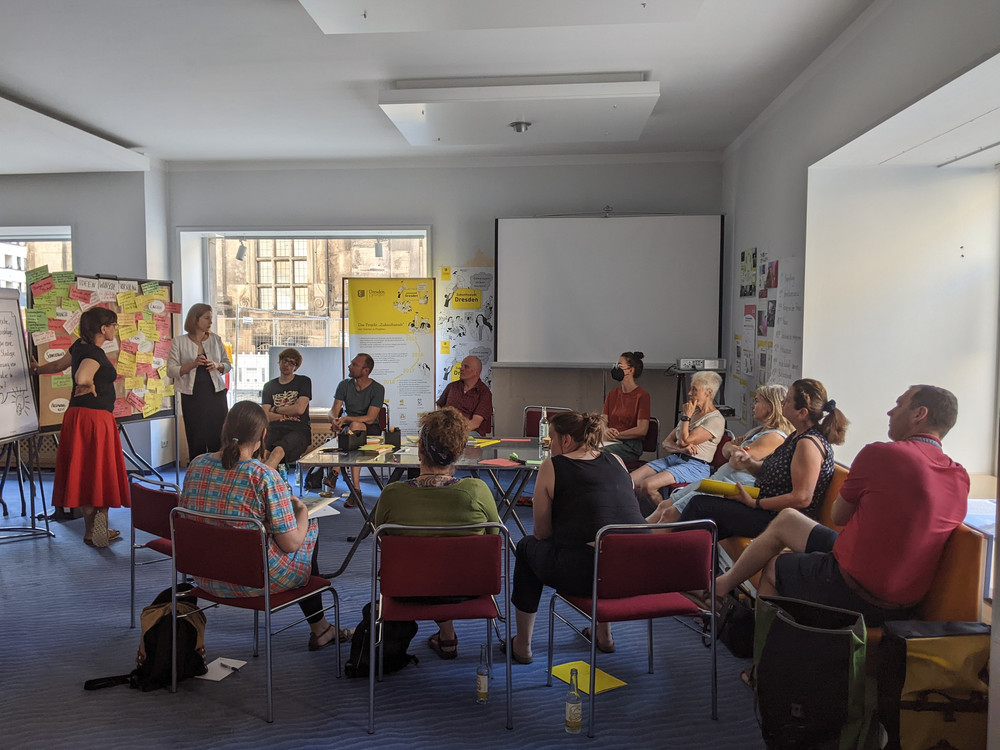 KI generiert: Das Bild zeigt eine Gruppe von Personen in einem Seminarraum, die an einer Diskussion oder einem Workshop teilnehmen. Im Vordergrund stehen zwei Frauen, eine davon präsentiert oder moderiert, während die anderen Teilnehmer an Tischen sitzen und zuhören.