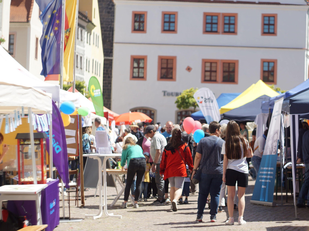 KI generiert: Das Bild zeigt einen belebten Marktplatz mit mehreren farbenfrohen Zelten und Ständen, an denen Menschen verschiedene Waren anbieten und besuchen. Der Hauptinhalt des Bildes ist ein lebhaftes Marktgeschehen mit vielen Besuchern und Ausstellern.