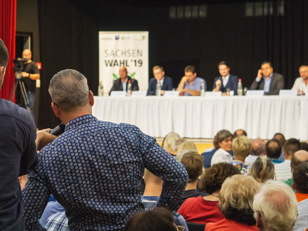KI generiert: Das Bild zeigt eine öffentliche Veranstaltung oder Podiumsdiskussion mit mehreren Teilnehmern auf der Bühne, die vor einem Publikum sprechen. Im Hintergrund ist ein Banner mit der Aufschrift "SACHSEN WAHL '19" zu sehen, was darauf hindeutet, dass es sich um eine Wahlveranstaltung in Sachsen handelt.