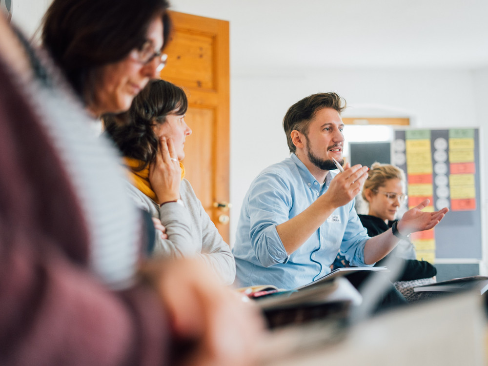 KI generiert: Das Bild zeigt eine Gruppe von Personen in einem Seminar- oder Meetingraum, wobei eine Person im Vordergrund gestikulierend spricht, während die anderen zuhören. Der Hauptinhalt des Bildes ist eine interaktive Diskussion oder Präsentation in einem professionellen oder pädagogischen Umfeld.