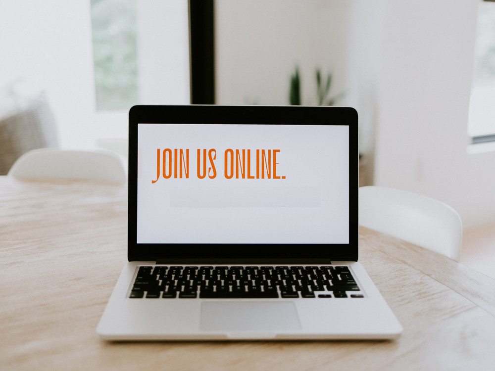 KI generiert: Das Bild zeigt einen Laptop auf einem Holztisch mit der Bildschirmanzeige "JOIN US ONLINE." Im Hintergrund sind unscharf zwei weiße Stühle zu erkennen. Der Hauptinhalt des Bildes ist die Einladung, online teilzunehmen.