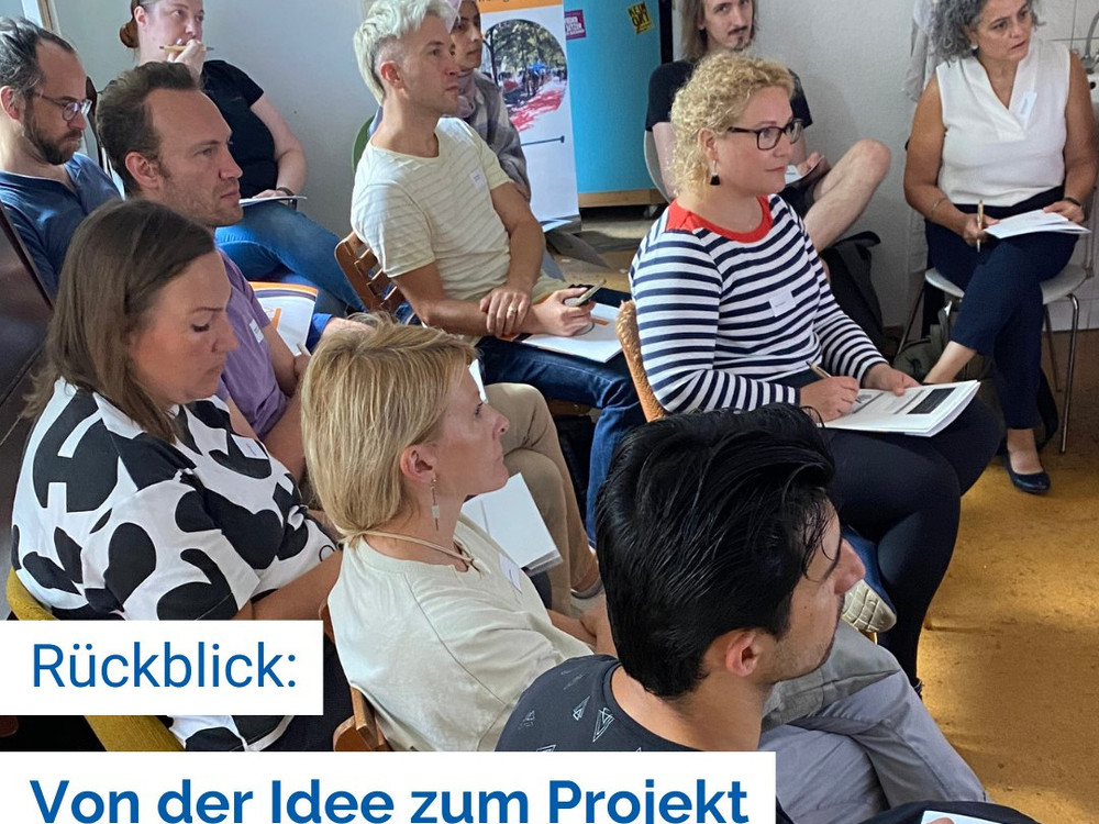 KI generiert: Das Bild zeigt eine Gruppe von Personen im Freien, die sich um eine Frau versammelt haben, die ein Blatt Papier hält und anscheinend etwas vorliest oder erklärt. Der Hauptinhalt des Bildes ist eine informelle Zusammenkunft oder Diskussion in einem Garten oder Park.