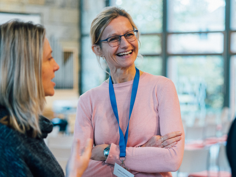 KI generiert: Das Bild zeigt zwei Frauen in einem hellen, modernen Innenraum, die sich angeregt unterhalten. Eine der Frauen trägt ein rosafarbenes Oberteil und eine blaue Namensschilder-Schnur um den Hals.